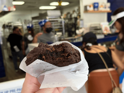 Un cookie au chocolat pâtissier, mis en avant devant la cuisine de Levain Bakery, animée par des clients.