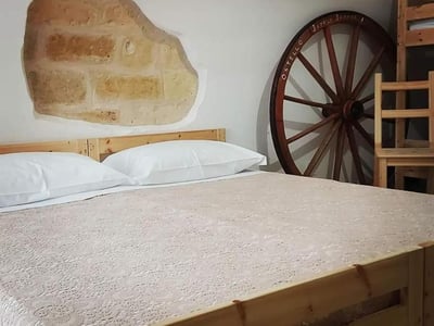 Cozy room at Japp'ca, comfortable bed with lace sheets, stone wall and decorative wheel in the background.