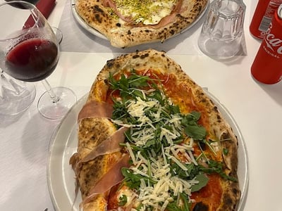 Artisanal pizza topped with prosciutto and arugula, accompanied by two glasses of red wine at Pizzeria Impasto Napoletano.