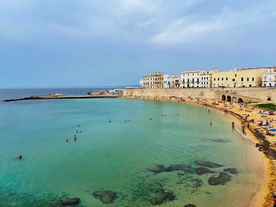 Serene beach of Gallipoli with bathers and historic buildings in the background under a cloudy sky.