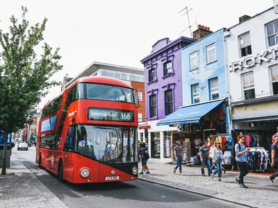Red double-decker bus traveling through the bustling streets of Camden Town, near colorful shops and passersby.