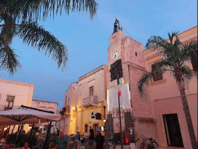 Tour charmant de Maruggio avec son horloge historique illuminée au crépuscule, entourée de palmier et d'animations locales.