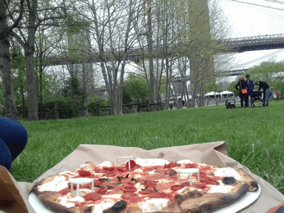 Pepperoni pizza from Grimaldi's Pizzeria with the Brooklyn Bridge in the background, on a green lawn.