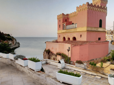 View of the pink castle in Tricase overlooking the sea, with plants in pots along the promenade. Peaceful atmosphere at dusk.