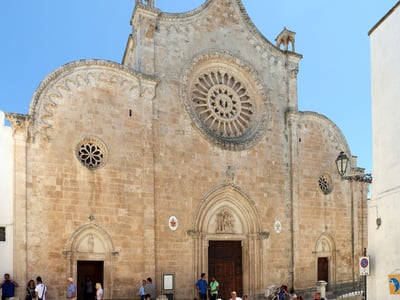 Sculpted façade of the Duomo di Santa Maria Assunta, surrounded by visitors, under a sunny sky in Ostuni, Italy.