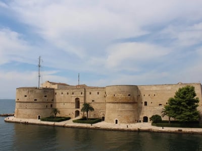 Historical fortifications of Taranto reflecting medieval architecture by the water, surrounded by palm trees.