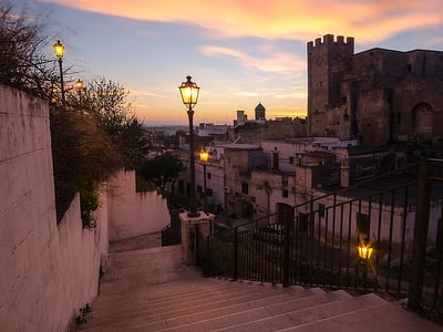 Illuminated stairs leading to the Castello Episcopio, surrounded by historic buildings under a colorful sky at dusk.
