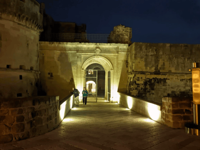 Une scène nocturne du Château d'Acaya, mettant en valeur l'entrée illuminée et des visiteurs admirant l'architecture historique.