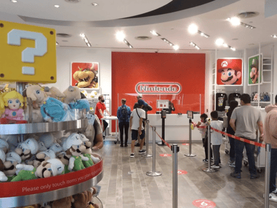 Magasin Nintendo animé avec des clients en file d'attente, peluches en promotion et décor coloré à Nintendo Store.