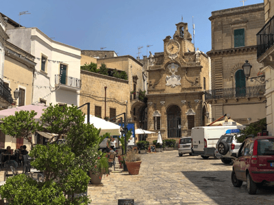 An animated view of Piazza Manfredi, with outdoor cafes and a historic building in thebackground.
