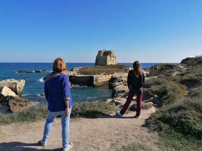 Two women admire the historic tower at Roca Vecchia, with a clear blue sea in the background and a sunny sky.