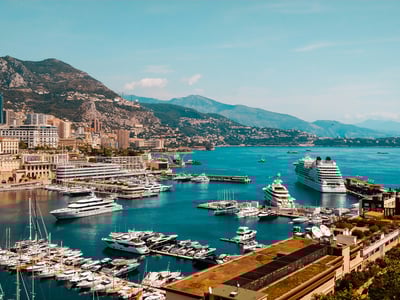 Panoramic view of the port of Monaco with luxurious yachts and the mountains in the background under a sunny sky.