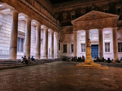 Belle cour intérieure du Convitto Palmieri, illuminée la nuit, avec des colonnes majestueuses et des visiteurs assis autour.