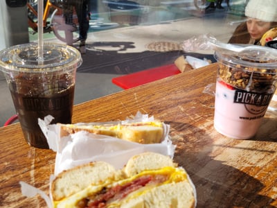 Lunch at Pick a Bagel with a hearty bagel, an iced coffee, and a fruit yogurt on display, urban atmosphere.