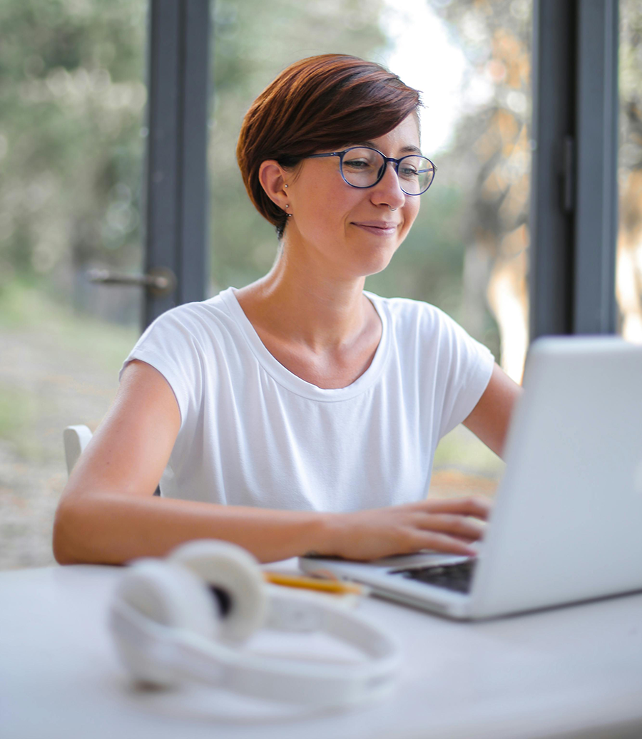 File Database and Email Backups Woman with short brown hair and glasses smiles while working on a laptop at a desk, with headphones and a pencil nearby, in a bright room with large windows.