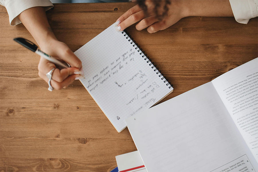 plan effectively A person writes notes in a notebook on a wooden desk beside an open book and some papers.