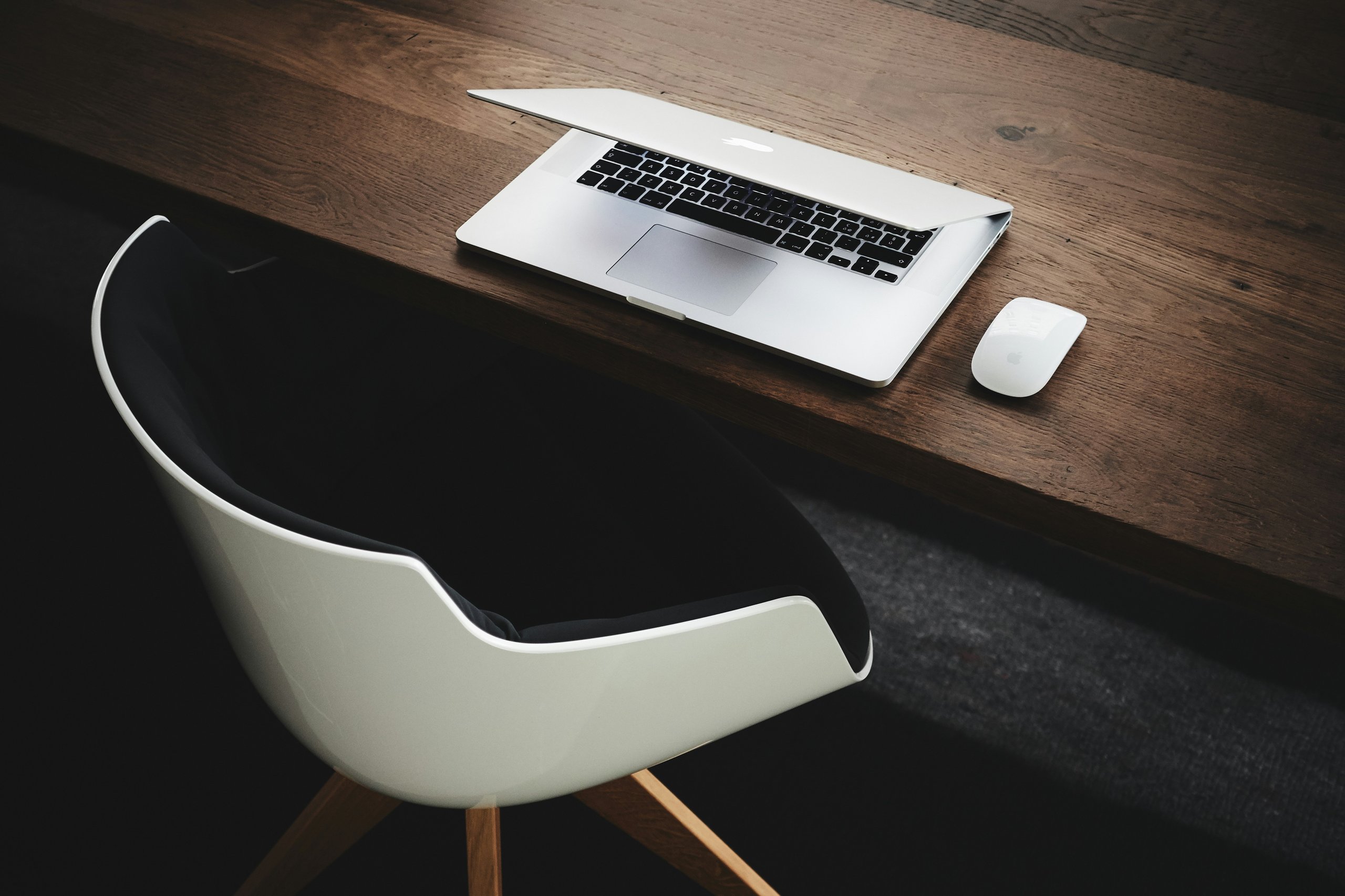 Update-WordPress A partially open laptop and a wireless mouse are on a wooden desk next to a modern black and white chair.