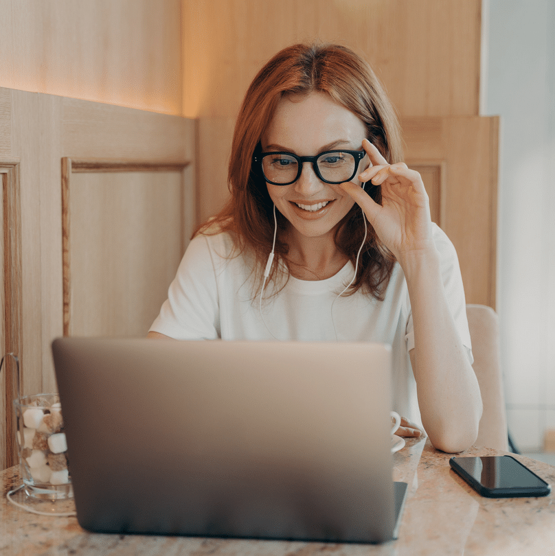 Woman with glasses smiling while using a laptop for domain registration at a home office setup.