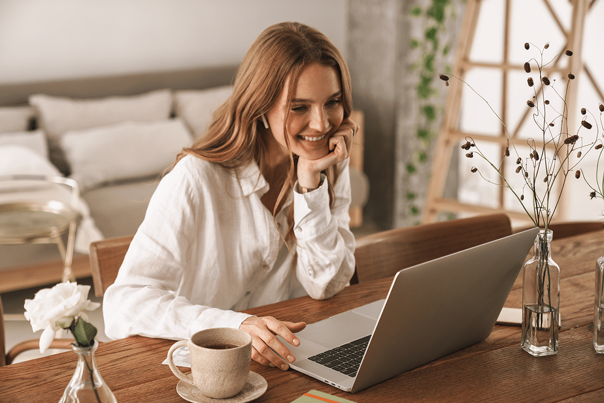 A woman with long hair smiles while using a laptop at a wooden table. She is wearing a white shirt and has a cup of coffee nearby. The background shows a cozy, decorated living space.