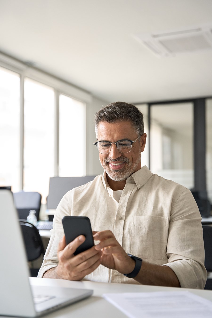 A man with glasses and a beard is smiling while looking at his phone. He is seated at a desk with a laptop in front of him. The background shows a modern office setting.