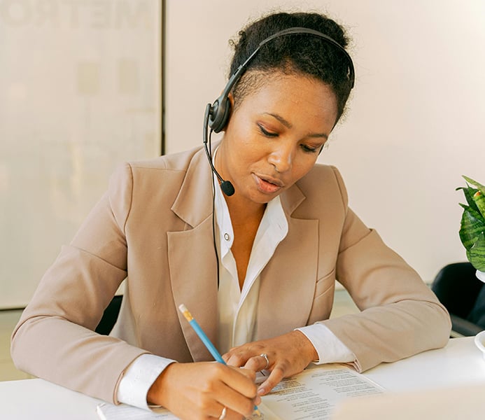 A person wearing a headset writes notes on a piece of paper while sitting at a desk.