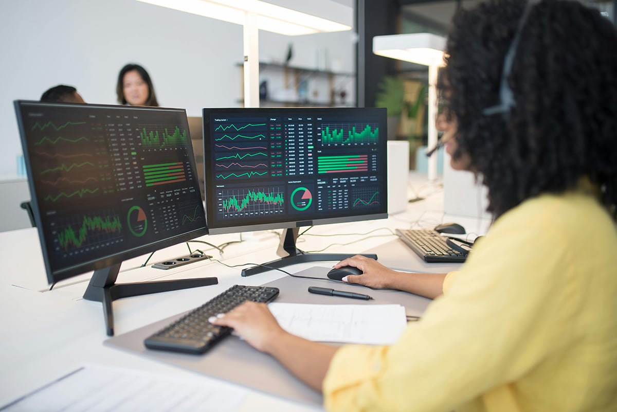 Person in a yellow shirt using two monitors displaying various graphs and charts, wearing a headset.