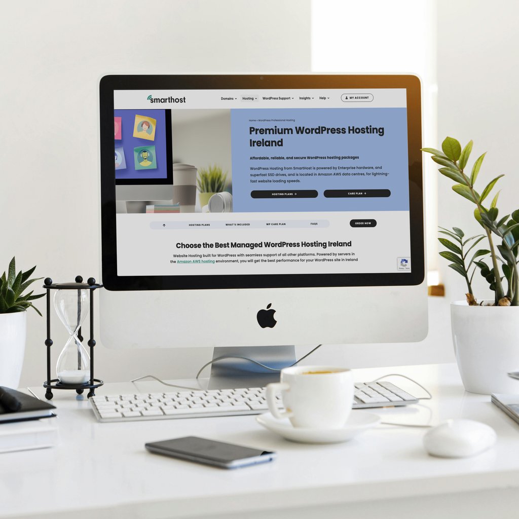 An iMac on a desk displays a website for premium WordPress hosting in Ireland. The workspace also features a keyboard, mouse, phone, hourglass, plant, and a cup—perfect for exploring top Web Hosting Ireland solutions.