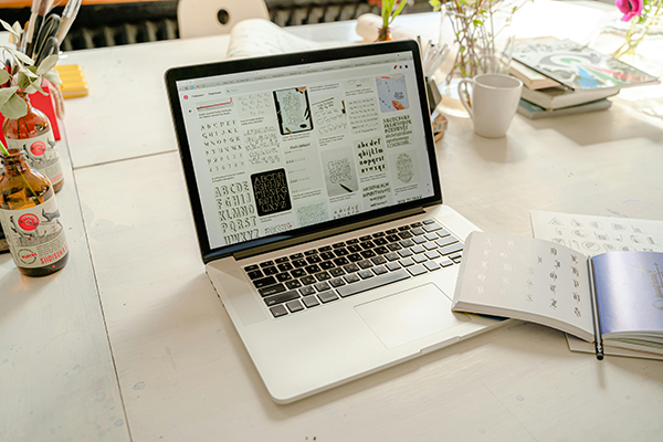 Optimise Font Usage and Delivery A laptop on a white table displays various fonts and designs. An open notebook, books, bottles, flowers, and a mug are also on the table.