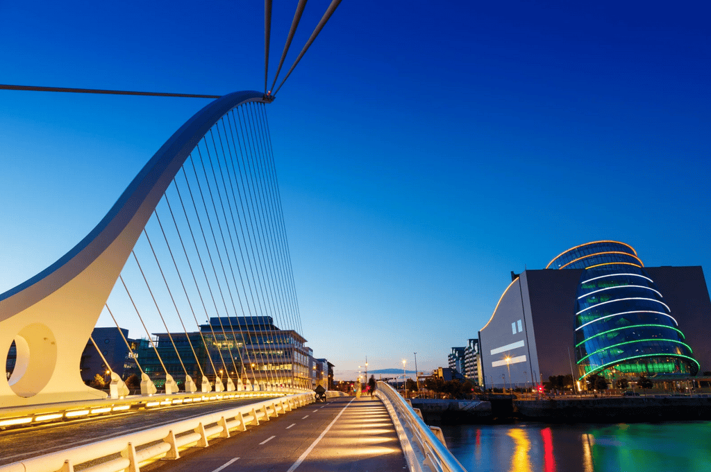 Benefits for Irish eCommerce Businesses A modern suspension bridge spans a river at dusk, with a curved glass building illuminated with colored lights on the right.