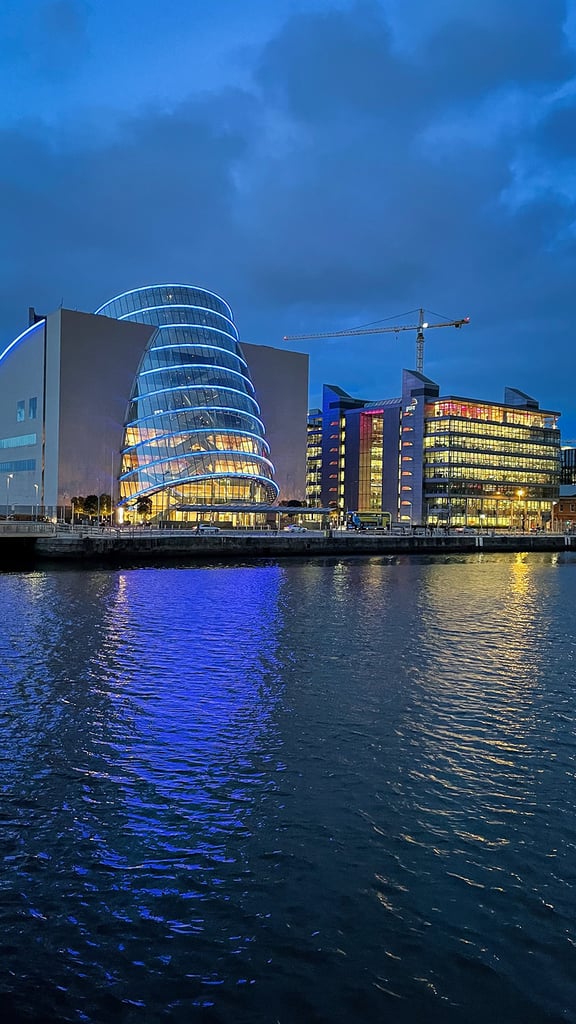 Modern glass buildings with bright lights reflecting on the water at dusk, under a cloudy blue sky. A crane is visible in the background.