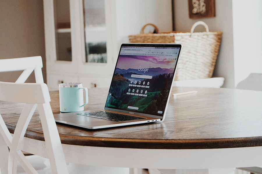 A laptop displaying a search engine homepage sits on a round wooden table next to a light blue mug in a bright, cozy room.