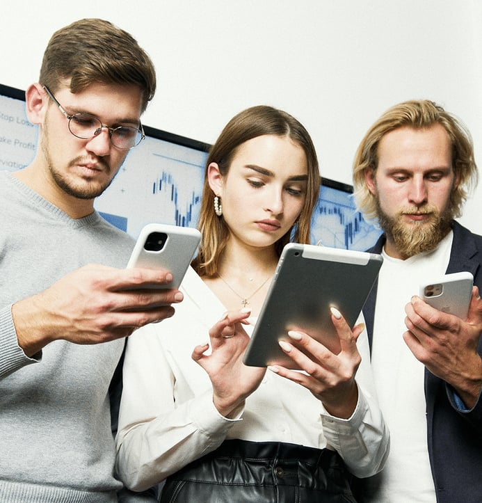 Three people standing close together, looking at electronic devices; two using smartphones and one using a tablet, with a digital screen in the background.