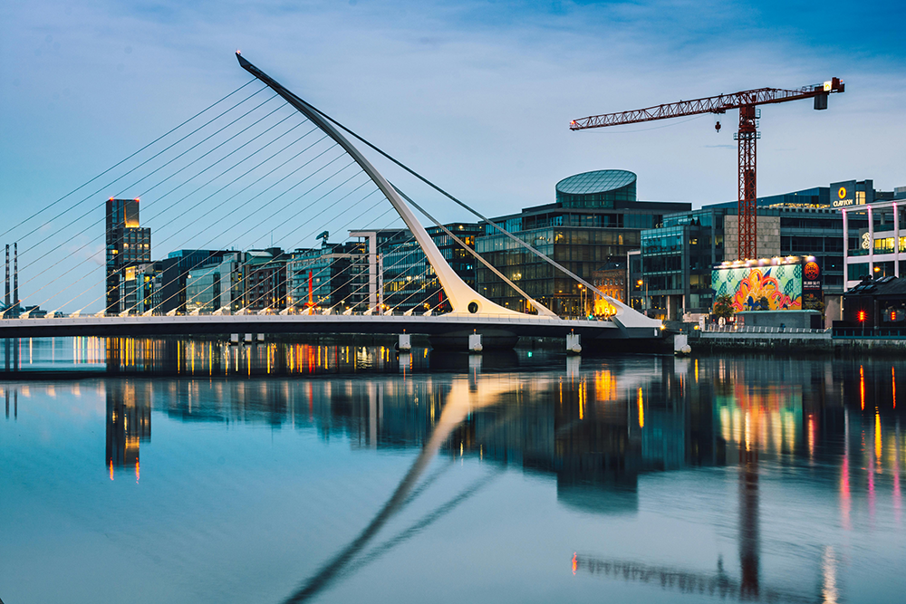 A modern cable-stayed bridge spans a calm river, with city buildings, a crane, and colorful mural visible in the background at dusk.