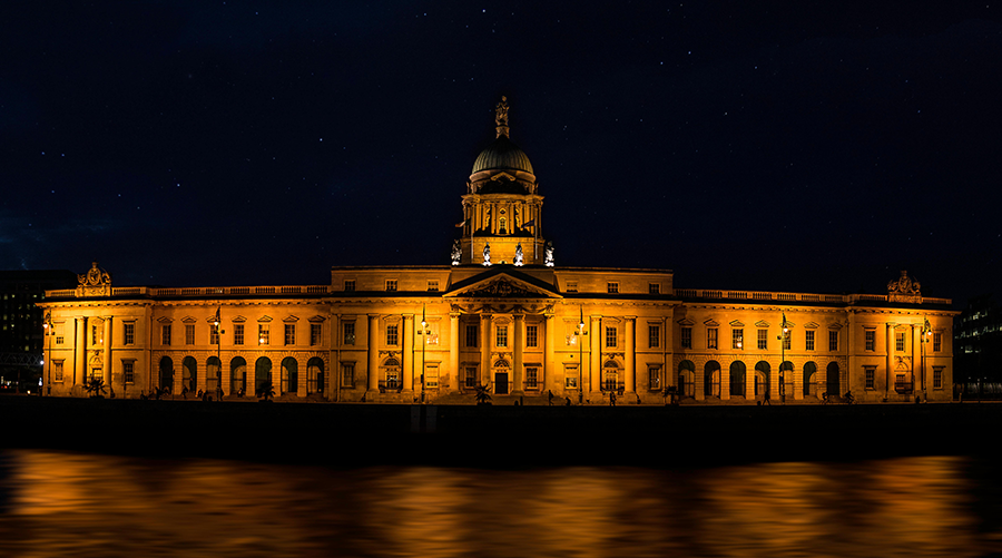 A large neoclassical building with a dome, illuminated by yellow lights at night, stands beside a calm body of water.