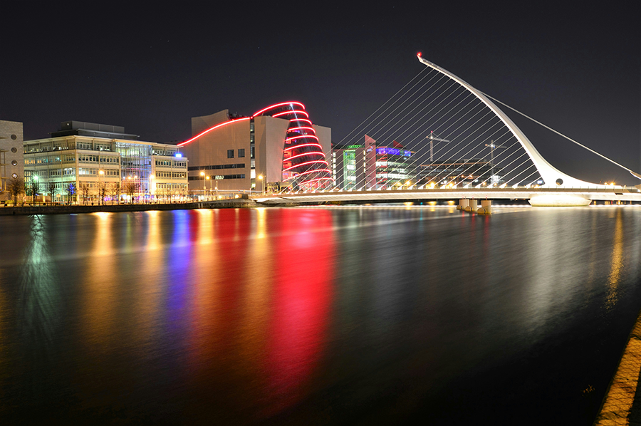 A night view of the Samuel Beckett Bridge and illuminated buildings reflecting on the River Liffey in Dublin, Ireland.