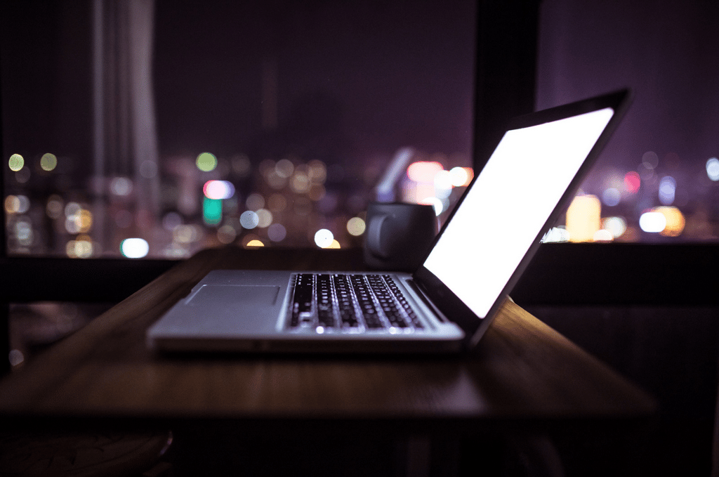 A laptop with a bright screen sits on a wooden table next to a mug, with city lights visible through a window in the background at night.