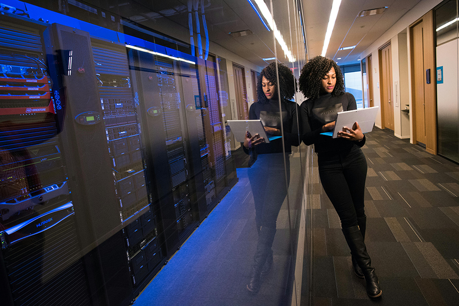 A woman stands in a hallway next to server racks, holding a laptop and looking at the screen, with her reflection visible in the glass wall beside her.