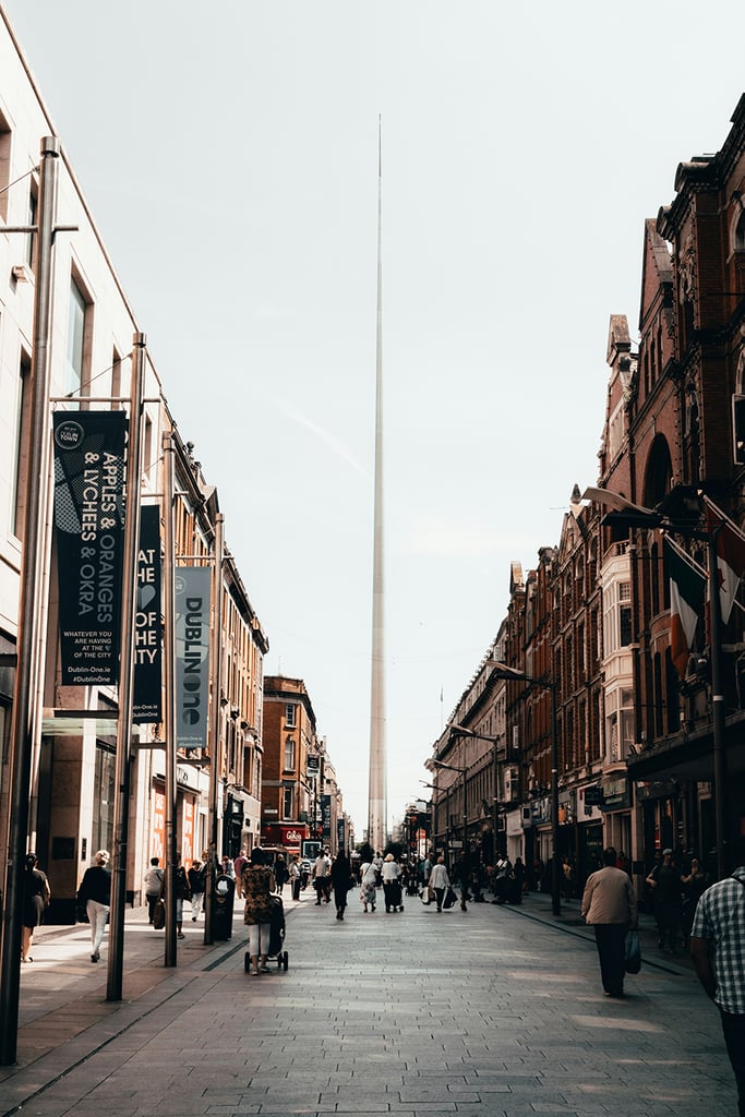 Why ISO 27001 hosting matters for SaaS products Pedestrians walk along a city street lined with shops and historic buildings, with a tall, pointed monument (the Spire of Dublin) rising in the center background.