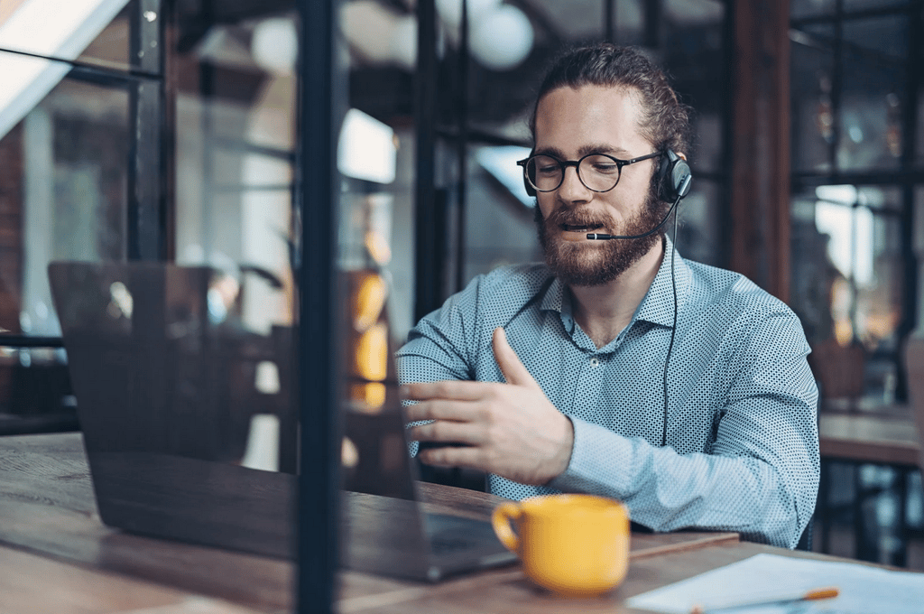 A man wearing a headset sits at a desk, talking during a video call on his laptop, with a yellow mug and a notebook nearby.
