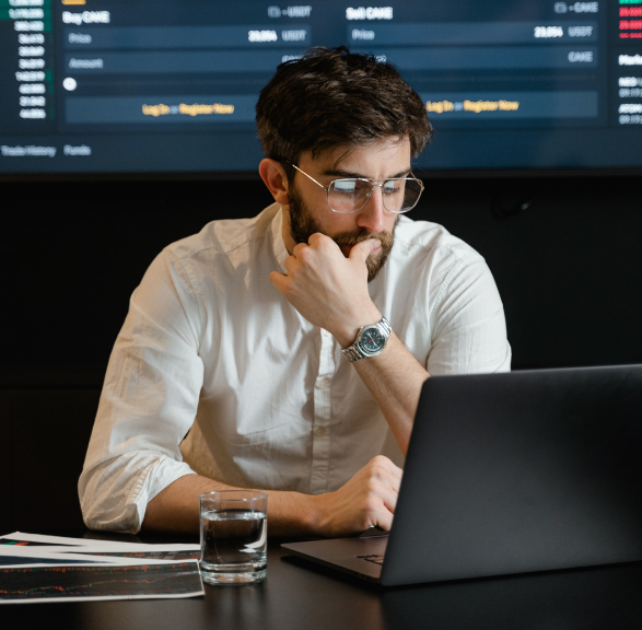 A man with glasses and a beard sits at a desk using a laptop, with financial charts on paper and a glass of water nearby; a trading screen is visible in the background.