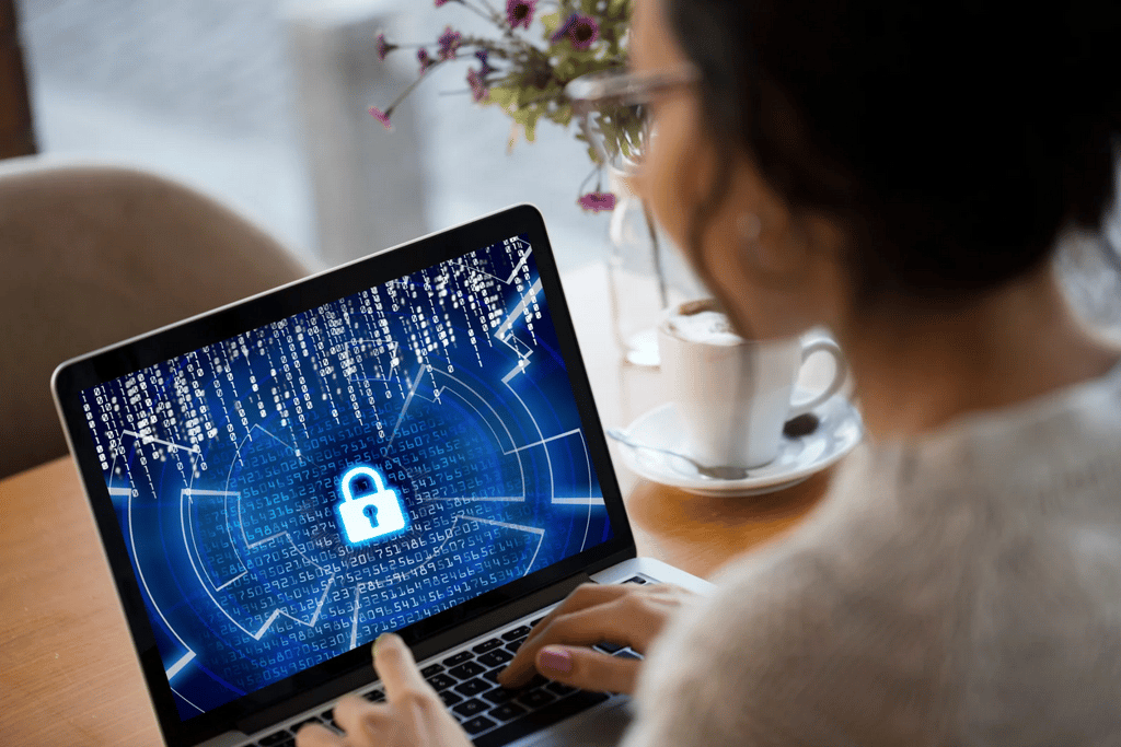 How ISO 27001 protects your website in the real world A person uses a laptop displaying a digital padlock and code, indicating cybersecurity or data protection, with a coffee cup and flowers in the background.