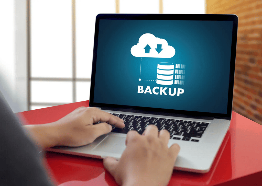 A person types on a laptop displaying a screen with a cloud backup icon and the word "BACKUP" in large letters.