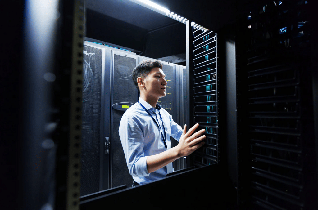 A man in business attire works inside a server room, standing between tall racks of computer servers and checking equipment.
