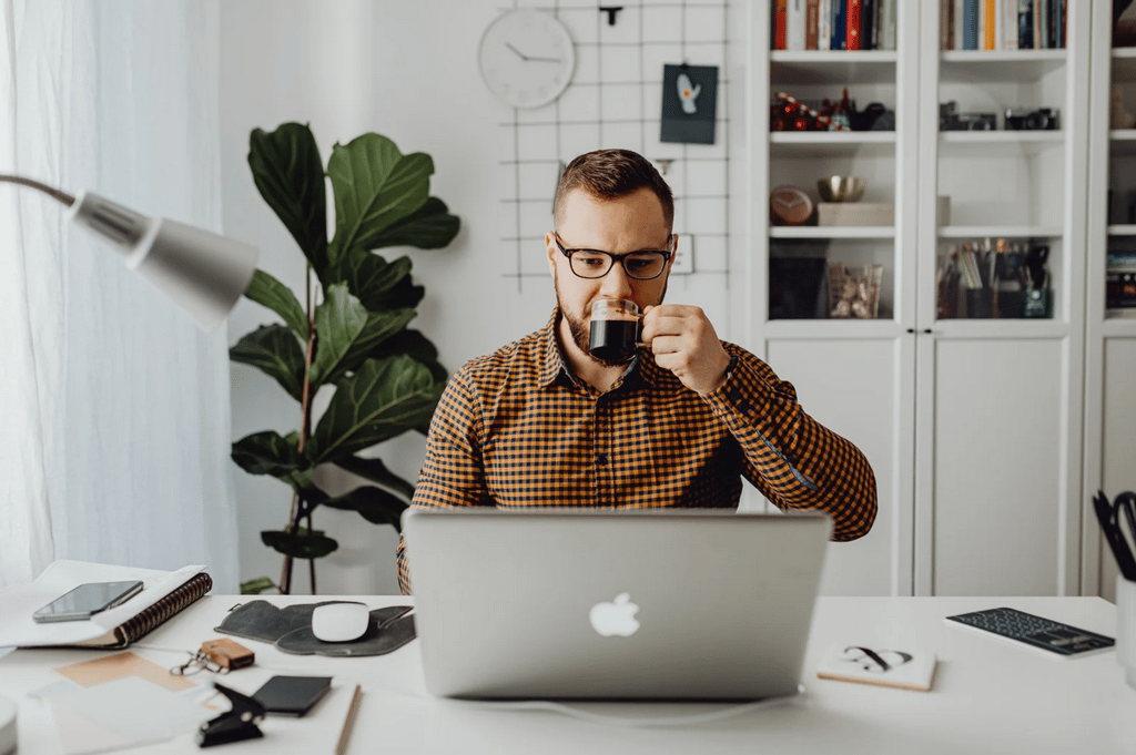 A man wearing glasses sits at a desk with a laptop, drinking from a mug in a home office with plants, books, and office supplies visible.