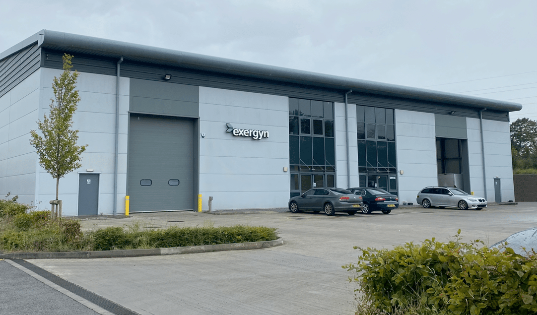 This image shows an industrial building with a large roller shutter door, glass entrance, a few parked cars, and the sign "Exergyn" on the facade.