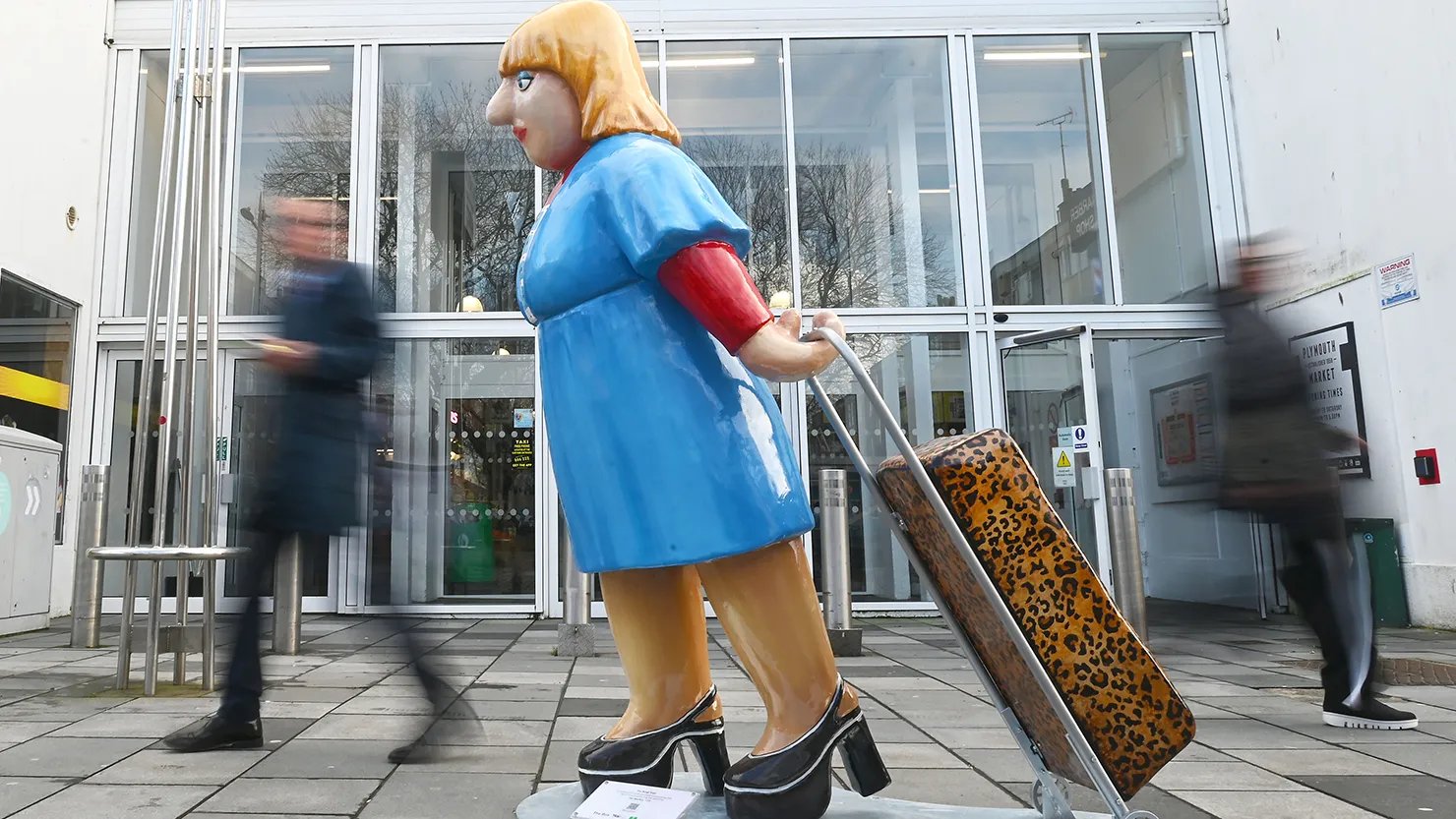 Photograph showing the sculpture of a woman in 1970s dress pulling a leopard print trolley in front of a market entrance