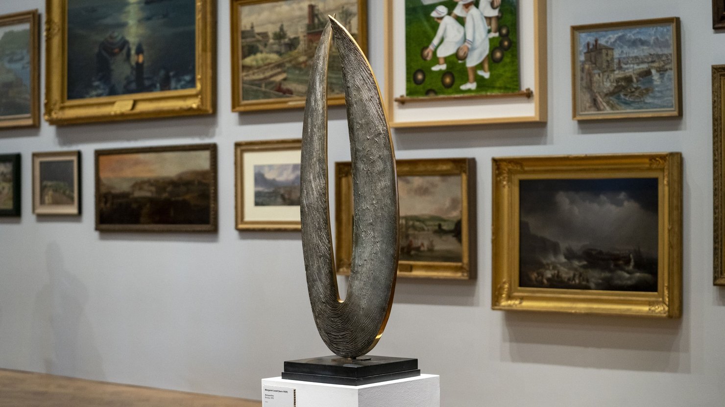 Photograph showing a bronze sculpture that appears like a sail, on a plinth within an art gallery.