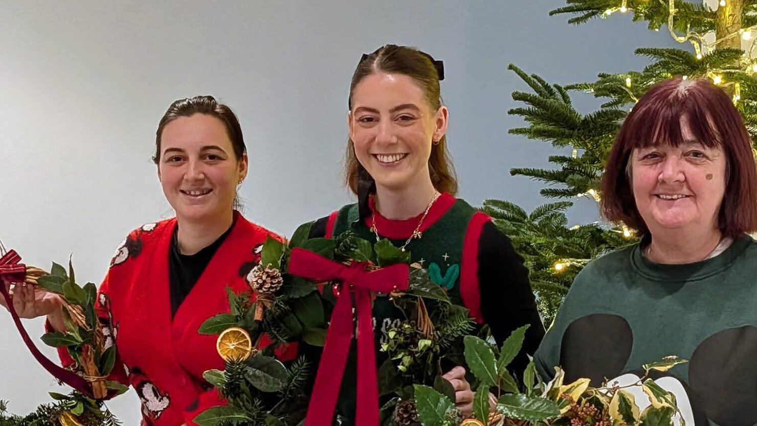 Three women wearing festive jumpers holding up Christmas Wreaths