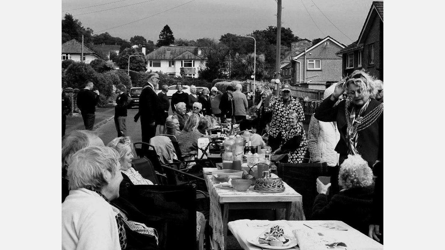 A black and white image of a group of people sat around a table at a street party enjoying food and drink