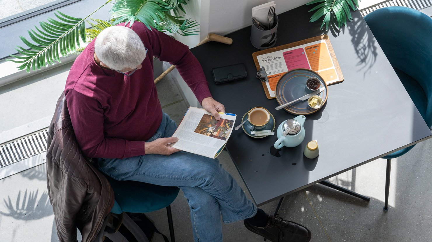 A man in a red shirt sits at a table reading a magazine and enjoying a pot of tea